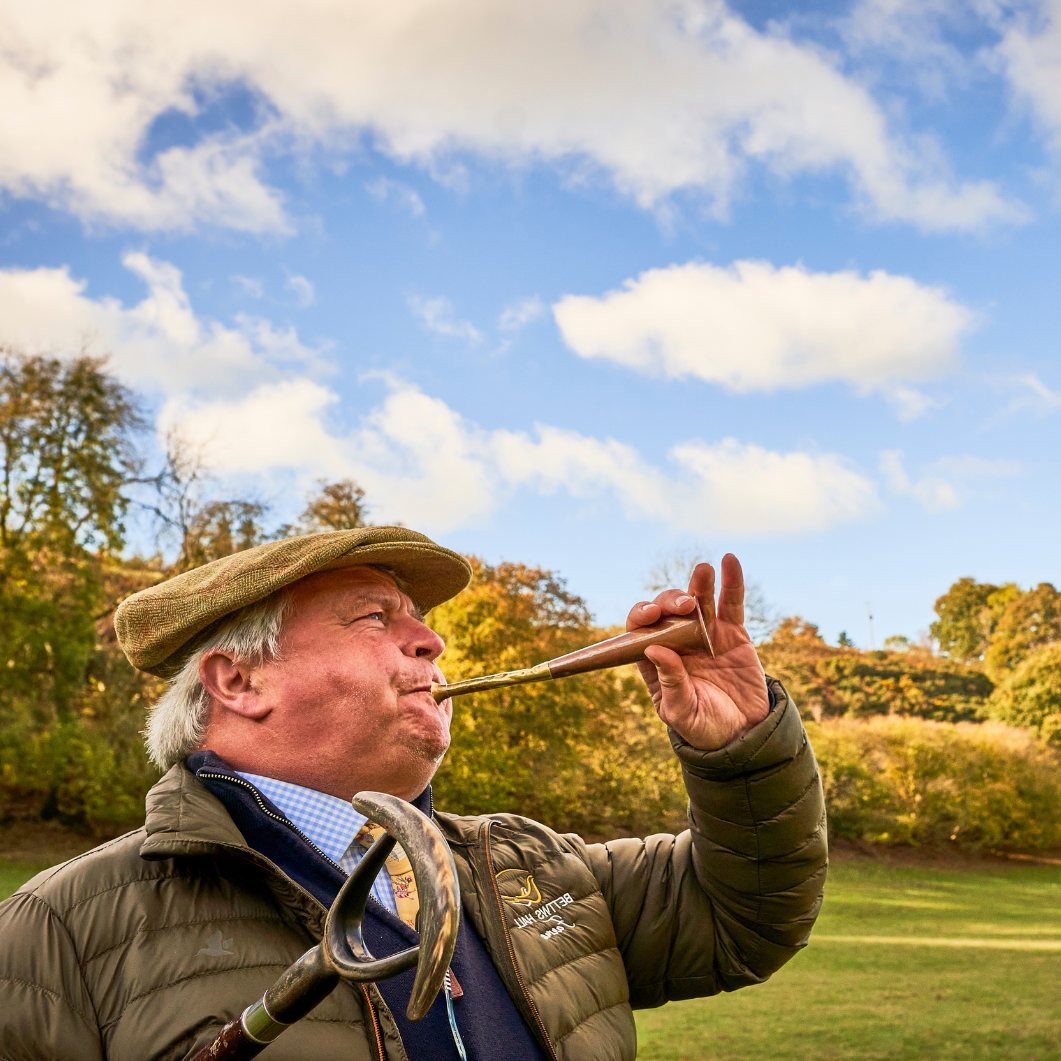 Man playing a hunting horn in a scenic outdoor setting with trees and blue sky.