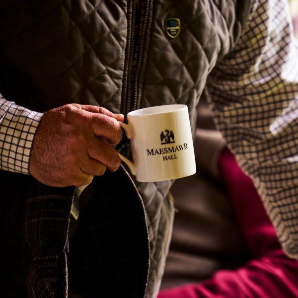 Person holding a white mug with 'Maesmawr Hall' branding, wearing a checkered shirt and plaid jacket.