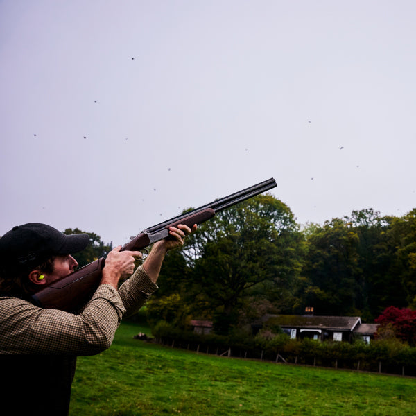 Person holding a shotgun in an outdoor setting with trees and a building in the background
