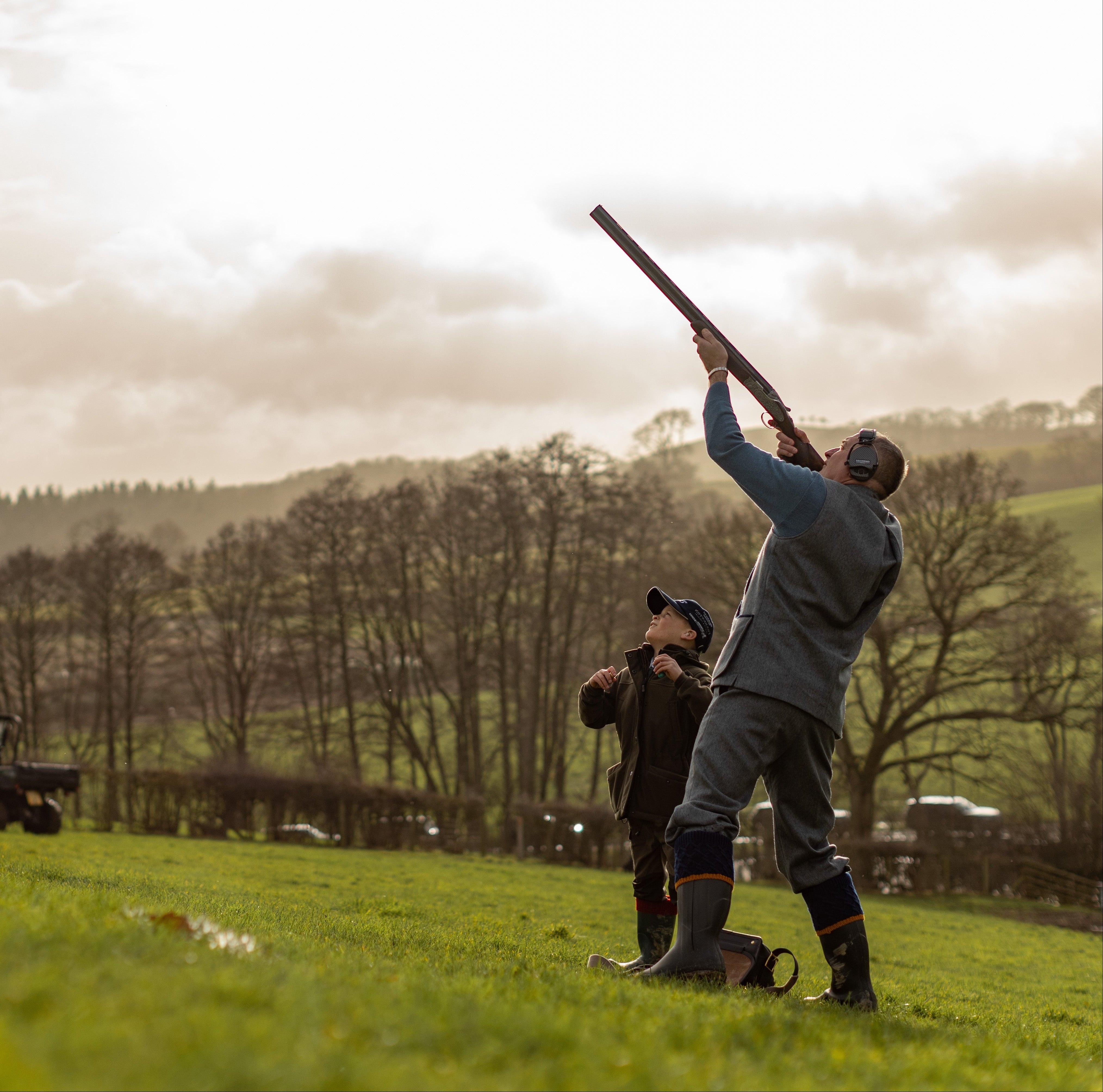 Man in a field with a shotgun, surrounded by trees and a cloudy sky.